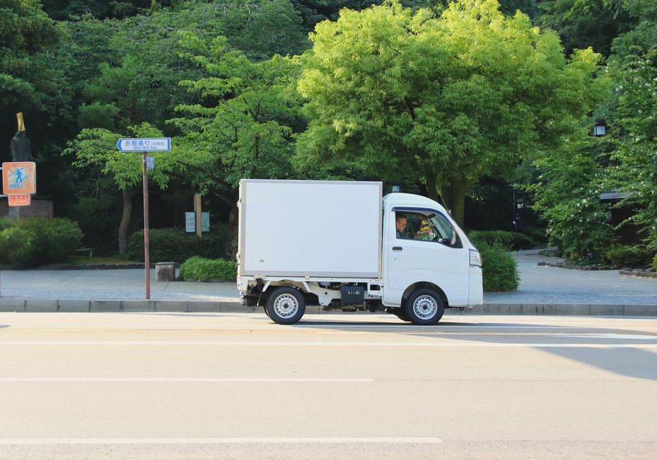 A city street scene with a mix of modern high-rise buildings and older structures, featuring various vehicles including a white van and a delivery truck parked and moving along the road. Overhead, mul