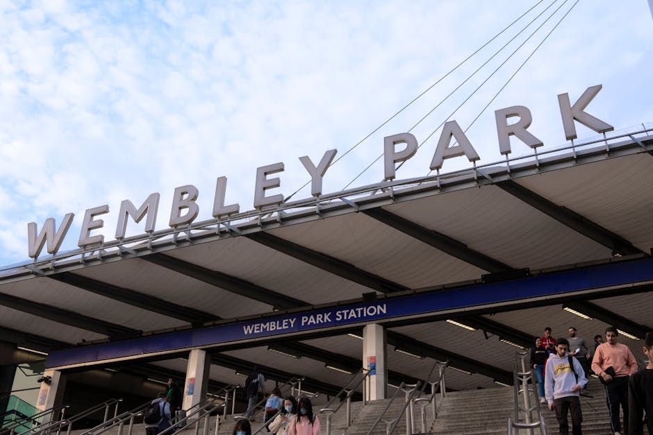 Photograph of Wembley Park Station's exterior showing the station's large metal sign spelling 'WEMBLEY PARK' mounted on the roof above the entrance. Below the sign, there is a blue and white station name board reading 'WEMBLEY PARK STATION'. The entrance features concrete stairs with metal handrails, with several commuters descending and ascending the steps, some carrying backpacks or bags. The station is situated outdoors under a partly cloudy sky, with visible overhead wires for electric trains. This scene captures the busy atmosphere of a travel hub, which is relevant to house removals and relocating services offered by Man with Van Tokyngton, illustrating the context of transportation logistics for home relocation and furniture transport.