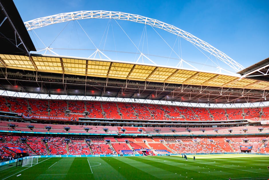 A panoramic view of a large, modern football stadium with a vivid green pitch, surrounded by multiple tiers of red seating. The partially covered roof extends over the seating area, with metal framework and overhead cables visible against a partly cloudy blue sky. Inside the stadium, there are no spectators or players, and the focus is on the empty seating and the pitch, which appears well-maintained. The ground level shows a loading area with several cardboard boxes, and a few movers in casual clothing are seen carrying and stacking items, preparing for a home relocation or furniture transport operation. The scene reflects the logistics involved in packing and moving, supported by equipment such as trolleys and blankets, indicative of a professional removal service, which could include [COMPANY_NAME], in the context of house removals or apartment moves near Wembley Stadium in Tokyngton, relevant to the page about best times to move in that area.