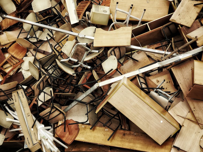 A large pile of disorganized discarded furniture and household items, including wooden tables, chairs with metal and wooden frames, plastic and fabric cushions, and various small objects, all stacked haphazardly inside a property or loading area. Some chairs are stacked upside down, while others are leaning against tables or lying on their sides. The scene also features a metal trolley and scattered packaging materials such as cardboard and plastic wraps. The lighting is natural, highlighting the clutter typical of a furniture clearance or home relocation process. This image illustrates the sorting and preparation phase involved in house removals, with Man with Van Tokyngton potentially assisting with furniture transport and packing services during residential moves or bulk waste collection, as noted on the Brent Council bulk waste rules page for Tokyngton moves.