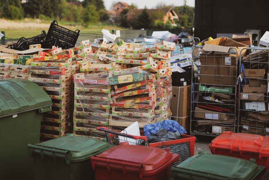 An outdoor area showing a large stack of cardboard boxes filled with fresh fruits, arranged on wooden pallets, with some boxes slightly open revealing contents, positioned next to a metal wire trolley loaded with various boxes and packaging materials, including brown paper and cardboard. Several plastic wheeled containers in green and red are placed nearby, some with lids, on a paved surface. In the background, a residential area with trees and houses is visible under daylight. The scene captures elements typical of home relocation or moving logistics, involving packing and loading items, with a focus on organizing boxes and packaging materials before transportation. The image reflects the logistical process of house removals, utilizing equipment such as stacking pallets, trolleys, and protective packaging, with the presence of a moving vehicle not visible in the frame. Man with Van Tokyngton occasionally manages such packing and furniture transport tasks as part of local house moves.