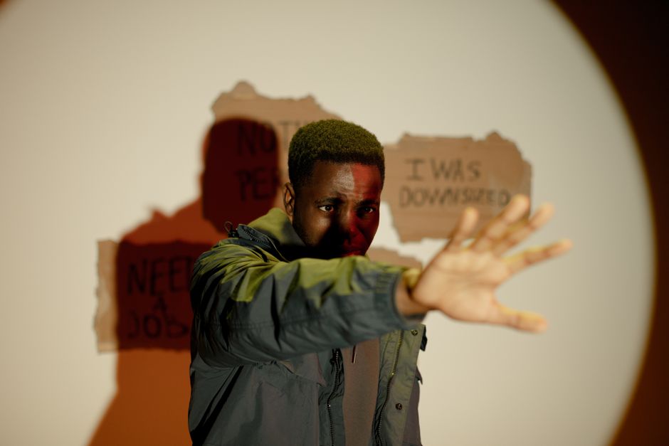 A young man with short, dark hair and dark skin stands indoors, extending his right arm forward with an open hand. He is wearing a green and grey jacket and appears to be reaching or gesturing towards the camera. Behind him, a torn piece of brown cardboard with visible text is taped to the wall, displaying partial phrases such as 'I was downsized,' 'Need a job,' and other words that are partially obscured. The lighting creates a shadow of the man on the wall, which highlights his figure and the text on the cardboard. The environment suggests an indoor setting related to house removals or relocation services, with the background and surroundings hinting at a scenario where the individual may be involved in a moving or packing process. Man with Van Tokyngton occasionally visible in the description, aligning with home relocation activities and moving logistics.