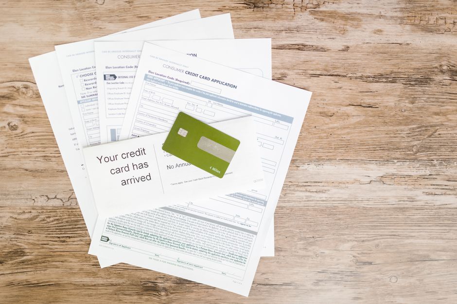 A set of four sheets of paper containing various forms, including a credit card application and a notice stating 'Your credit card has arrived,' are spread out on a light wooden surface. The papers are positioned in a slightly overlapping arrangement, with the top sheet displaying the message. A green plastic or cardboard credit card mock-up is placed on top of the forms, partially covering the text. The scene portrays paperwork typically associated with home relocation or moving preparations, suggesting activities related to financial arrangements or documentation for house removals services. The background is a plain, natural wood texture, and no other objects are visible. The image reflects the administrative aspects of house removals, such as managing finances and logistics, captured in a neutral and professional style, consistent with services like those offered by Man with Van Tokyngton.
