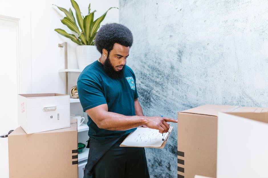 A man with dark curly hair, a full beard, and wearing a dark teal T-shirt, is standing indoors next to a stack of cardboard boxes, some sealed and others open, with packing materials visible around him. He is holding a clipboard with a pen and appears to be reviewing or checking a moving inventory list. The background includes a white shelf with a potted plant and other household items, and the wall behind him has a light, textured finish. The setting suggests a home environment during a house removal process, with furniture and boxes prepared for transport. The scene reflects the packing and moving logistics carried out by professionals such as Man with Van Tokyngton, supporting home relocation and furniture transport services.