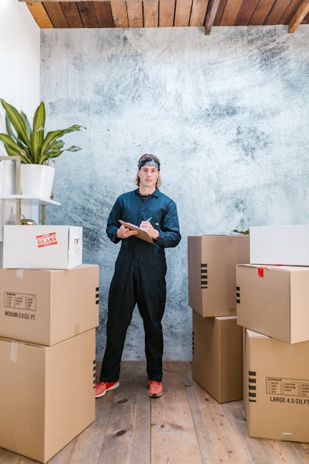 Inside an interior room with a wooden ceiling and a textured, light-colored wall, a woman dressed in navy work overalls, a knitted headband, and red shoes stands among several large cardboard boxes of various sizes. Some boxes are sealed with packing tape, and one has a label indicating a medium size with volume measurements. A white shelving unit in the corner holds a potted plant and other items. The woman is holding a clipboard and pen, likely reviewing a packing or home relocation checklist as part of a furniture transport or packing process. The floor is wooden, and the scene appears to be part of a house moving or professional removals service provided by Man with Van Tokyngton, supporting house removals and relocation logistics.