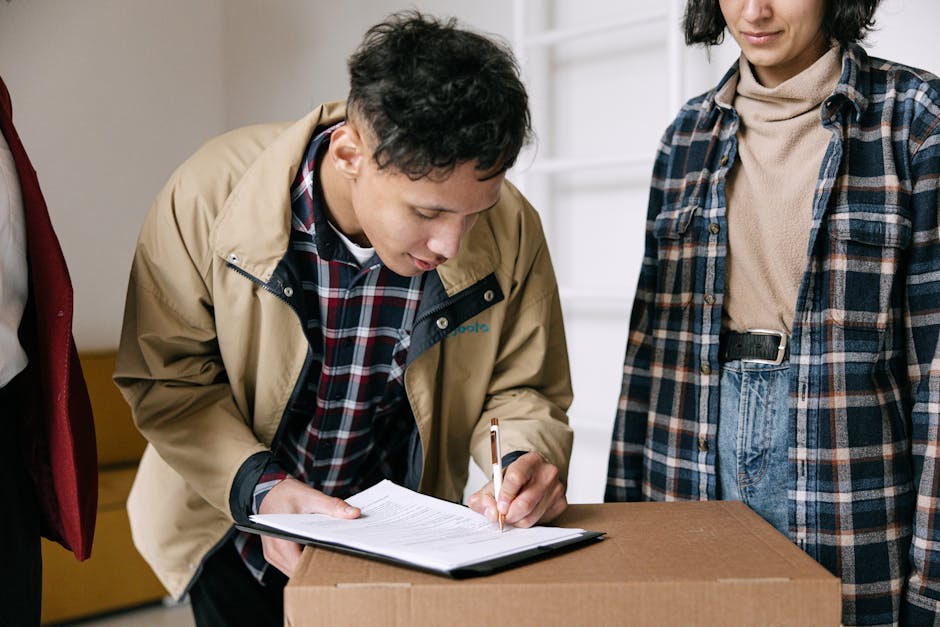 A man with dark curly hair, a full beard, and wearing a dark teal T-shirt, is standing indoors next to a stack of cardboard boxes, some sealed and others open, with packing materials visible around him. He is holding a clipboard with a pen and appears to be reviewing or checking a moving inventory list. The background includes a white shelf with a potted plant and other household items, and the wall behind him has a light, textured finish. The setting suggests a home environment during a house removal process, with furniture and boxes prepared for transport. The scene reflects the packing and moving logistics carried out by professionals such as Man with Van Tokyngton, supporting home relocation and furniture transport services.