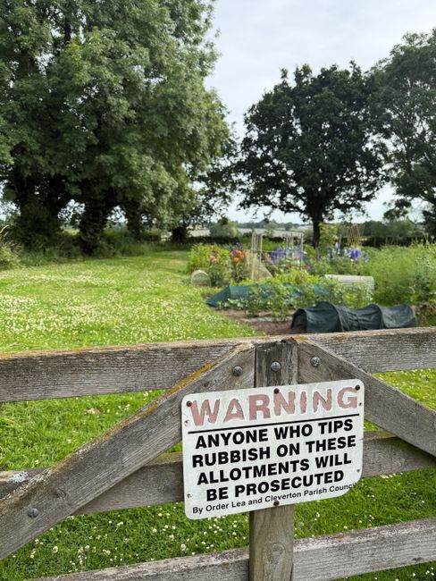 A close-up view of a weathered wooden fence with a white warning sign attached, situated in a rural outdoor setting. The sign has bold red and black text that reads, 'WARNING: Anyone who tips rubbish on these allotments will be prosecuted,' indicating strict rules against illegal dumping. Behind the fence, there is a garden area with various plants, raised flower beds, and gardening equipment, including black plastic seedling protectors and trellises, suggesting ongoing gardening or home relocation preparations. In the background, large green trees and open fields are visible under a cloudy sky, emphasizing a peaceful, countryside environment. The scene naturally reflects outdoor maintenance and community garden vigilance, aligning with the themes of property management and outdoor space care often associated with house removals and moving services.