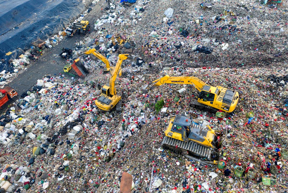 An aerial view of a large landfill site filled with scattered waste and rubbish, including plastic bottles, bags, and household debris. Multiple yellow excavators are actively engaged in moving and sorting through the waste materials atop mounds of trash. The scene captures machinery lifting and excavating heaps of waste within the site, with some pieces of rubbish being transferred into trucks or other containers. The environment appears open and exposed, with a mixture of natural and man-made materials visible, highlighting the scale of waste accumulation. This setting reflects the process of waste disposal and management, which can be related to waste removal services offered by companies such as Man with Van Tokyngton as part of comprehensive home or business relocation logistics involving disposal and recycling of unwanted items.