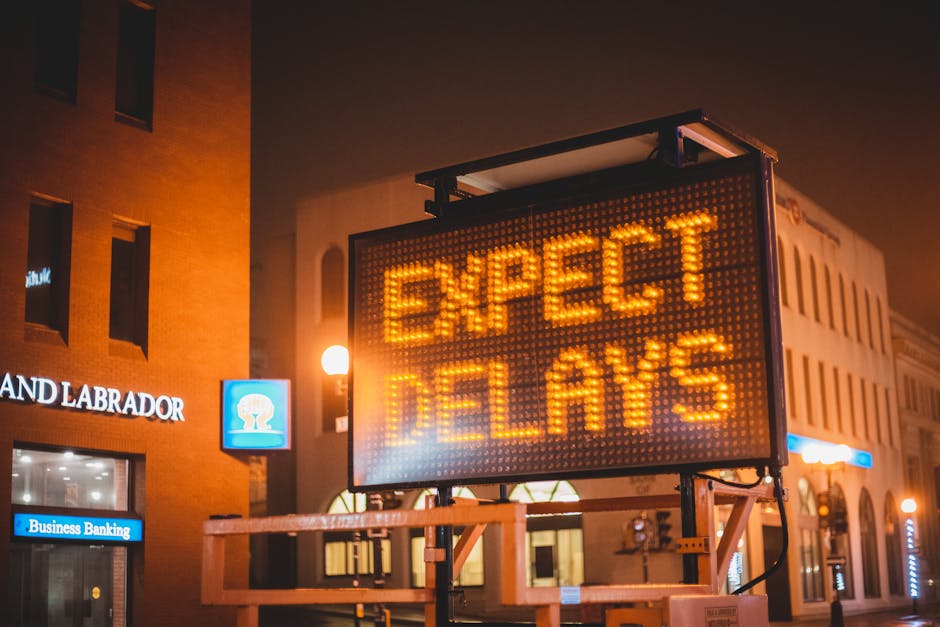 A large illuminated electronic traffic sign displays the message 'EXPECT DELAYS' in bright amber lights, mounted on a metal frame situated on the pavement outside a multi-storey building at night. The