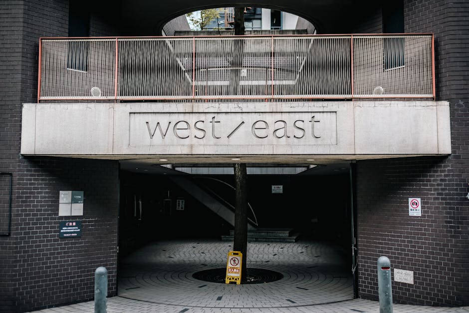 The image shows the underside of a modern building with a concrete sign reading 'west / east' mounted on the wall beneath a balcony with a copper-colored metal railing. The entrance area features an o